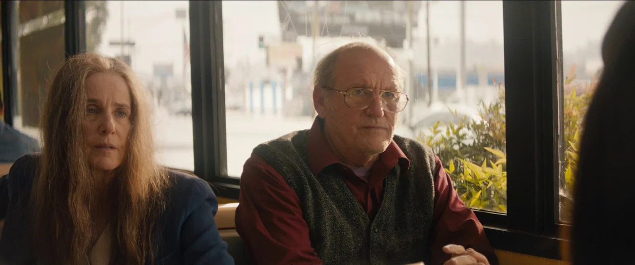 Kajillionaire (2020) movie still: medium — A window booth in a daytime setting with a blurred urban background visible outside. A man in a sweater…