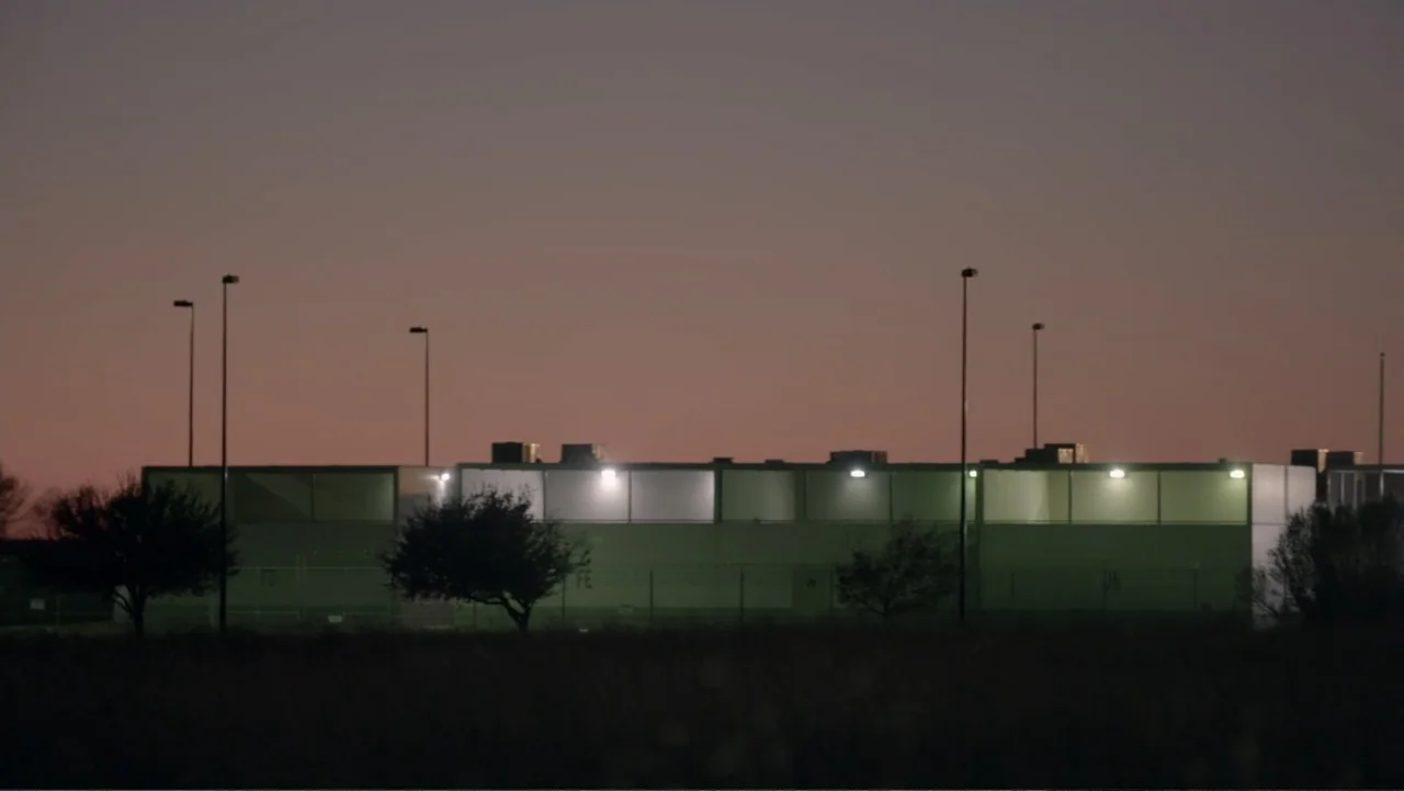 Torn Apart: Separated at the Border (2019) movie still: establishing — A low concrete building with fenced enclosures stands in a dark, empty field.…