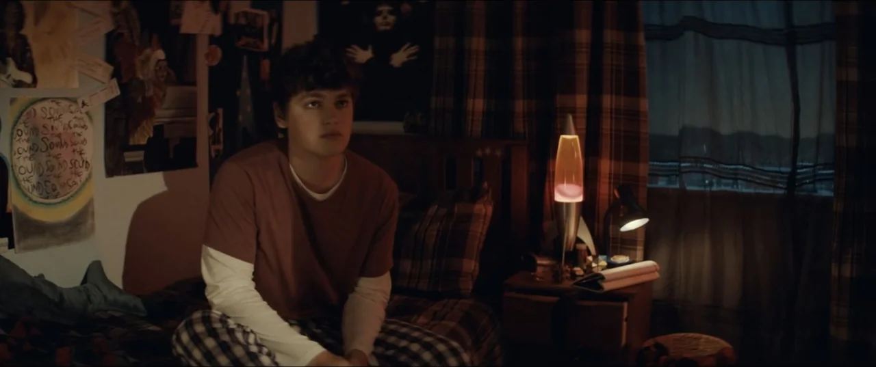 Bonus Track (2024) movie still: medium — A teenage bedroom contains plaid bedding, wall posters, and a glowing lava lamp on the nightstand. The male…