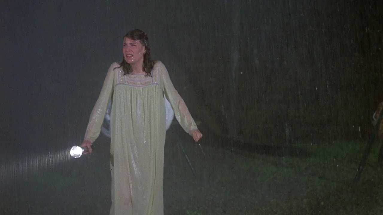 Friday the 13th (1980) movie still: fullbody — A dark, rainy outdoor area at night with heavy downpour visible in the frame. A young woman in a…