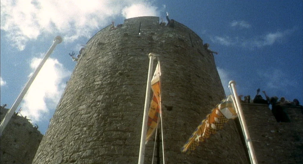 Jabberwocky (1977) movie still: wide — A massive, cylindrical stone castle tower rises against a bright blue, partly cloudy sky. Several heraldic…