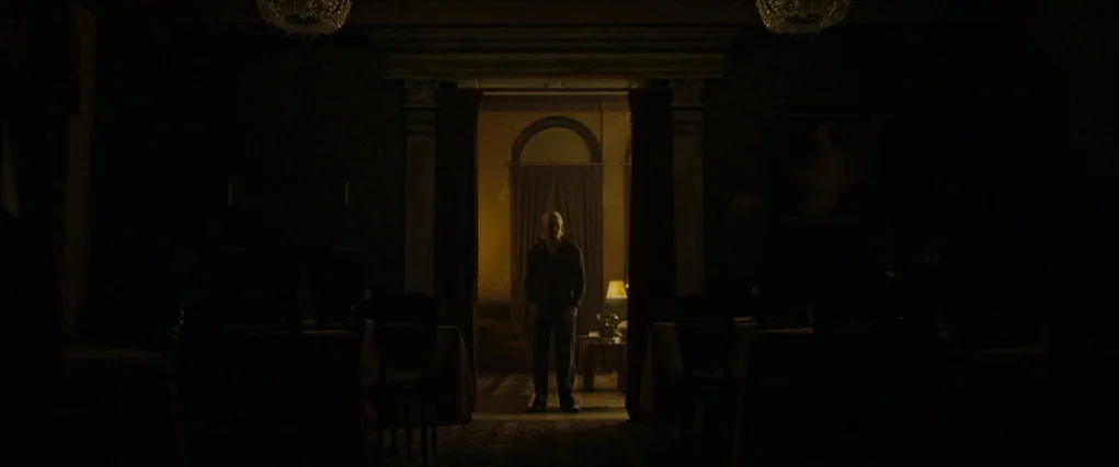 The Curious Case Of Benjamin Button (2008) movie still: wide — A dimly lit, large hall with dark furniture and two prominent overhead chandeliers.…