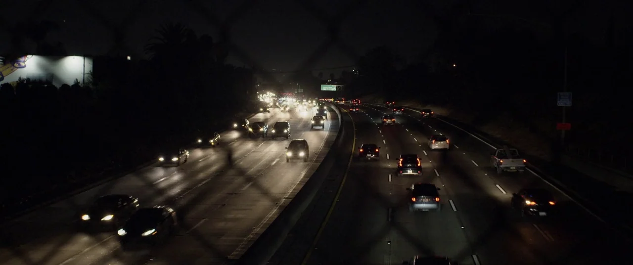 Blindfire (2020) movie still: wide — A multi-lane highway at night viewed through a chain-link fence. Numerous cars move along the road with their…