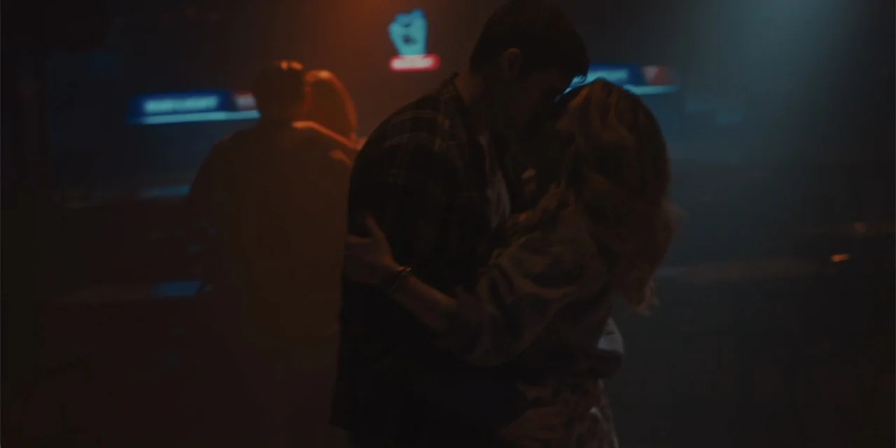 All Roads to Pearla (2019) movie still: medium — A dimly lit bar interior contains glowing neon signs and silhouettes of other patrons. A man and a…