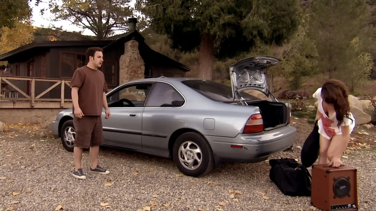 Hot Gimmick: Girl Meets Boy (2019) movie still: wide — A gravel driveway sits before a brown wooden cabin with a stone chimney. A man and woman…