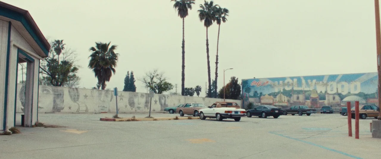 MaXXXine (2024) movie still: wide — An open asphalt parking lot sits beneath an overcast sky with tall palm trees. A long wall displays a faded…