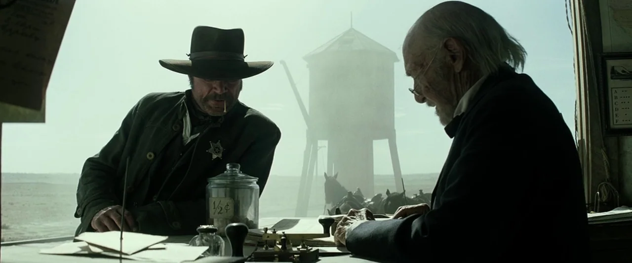 The Lone Ranger (2013) movie still: medium — A dusty, sunlit telegraph office with a view of a water tower outside. A sheriff stands opposite an…