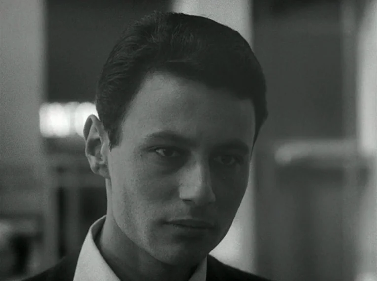 Le Petit Soldat (1963) movie still: closeup — A close-up view shows a young man's head and shoulders against a blurry background. He gazes slightly…