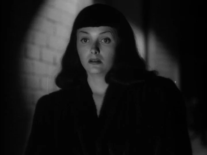 The Seventh Victim (1943) movie still: closeup — A woman stands in front of a textured brick wall at night. Strong light casts deep shadows across…