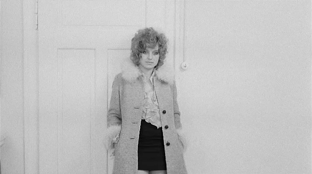 Love Is Colder Than Death (1969) movie still: medium — A woman stands against a white door in a minimally furnished room. The background wall is…