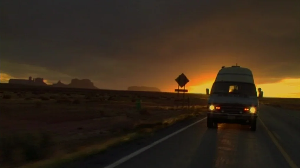 Land of Plenty (2004) movie still: wide — A van drives on a desolate highway through a vast desert landscape at sunset. Silhouetted rock formations…