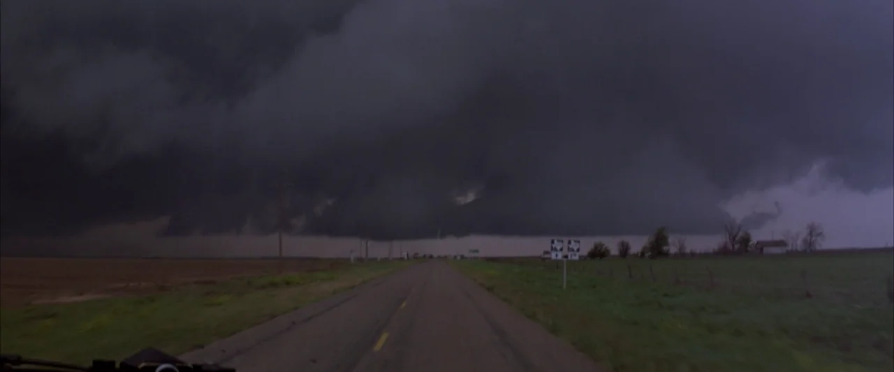 Twister (1996) movie still: wide — A desolate paved road stretches forward through a rural landscape under massive, dark storm clouds. Low, thick…