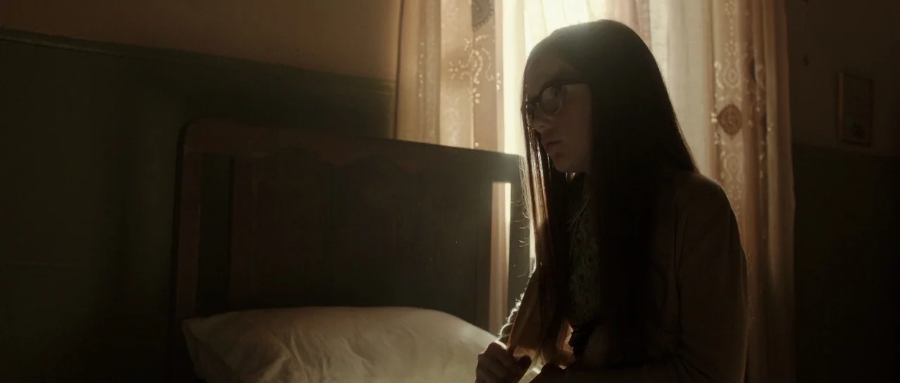 Predestination (2014) movie still: medium — A dimly lit room contains a bed with a dark headboard and curtains. A teenage girl with glasses sits…