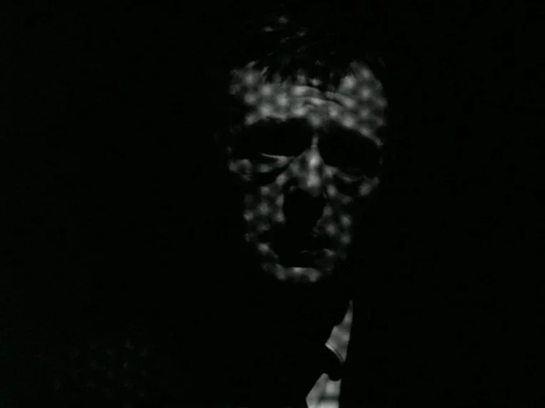 Death and Transfiguration (1983) movie still: closeup — A man's face is illuminated by light passing through a perforated surface. He stands in…
