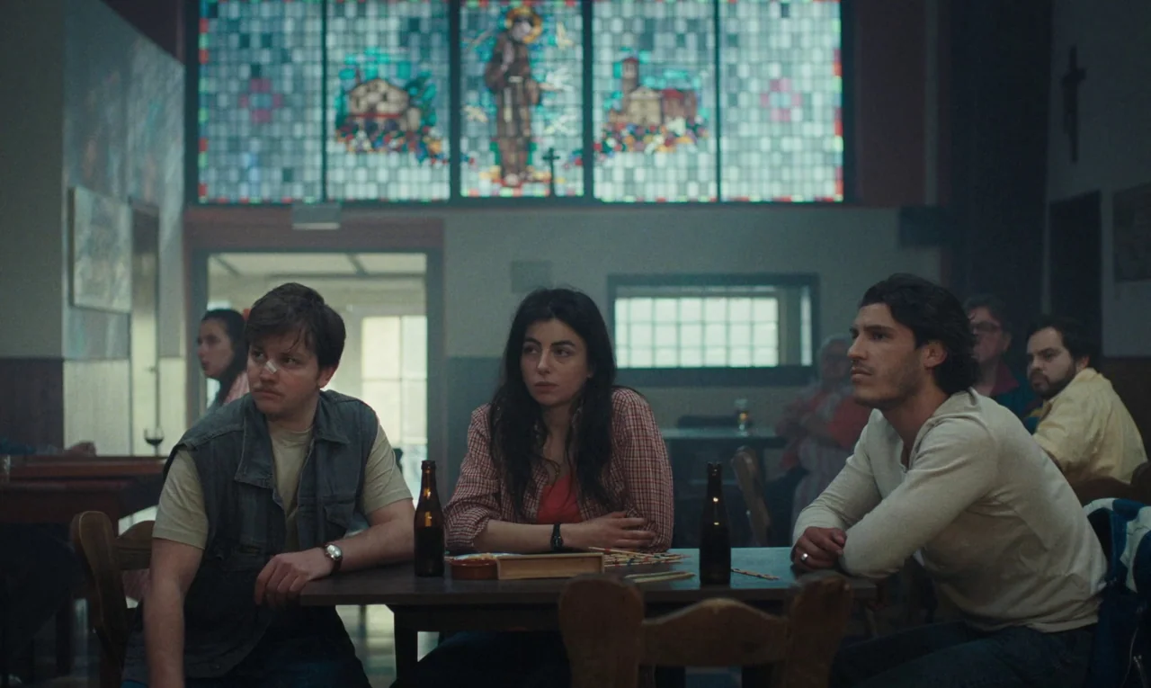 Maldoror (2024) movie still: medium — A community room features a prominent stained glass window depicting a saint at the back. Three young adults…
