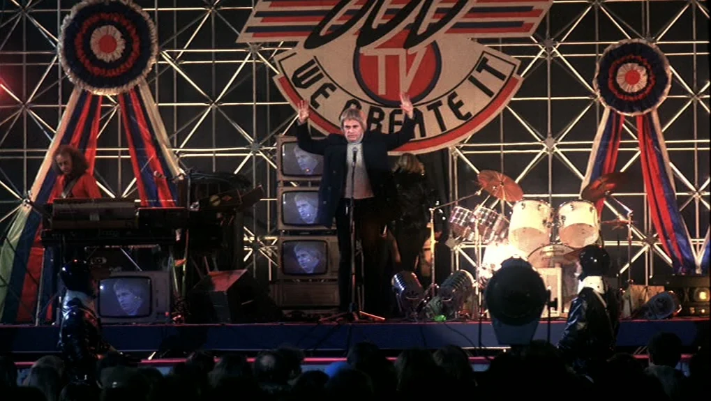 The War of the Worlds: Next Century (1981) movie still: wide — A brightly lit concert stage features a geometric metal grid backdrop with large…