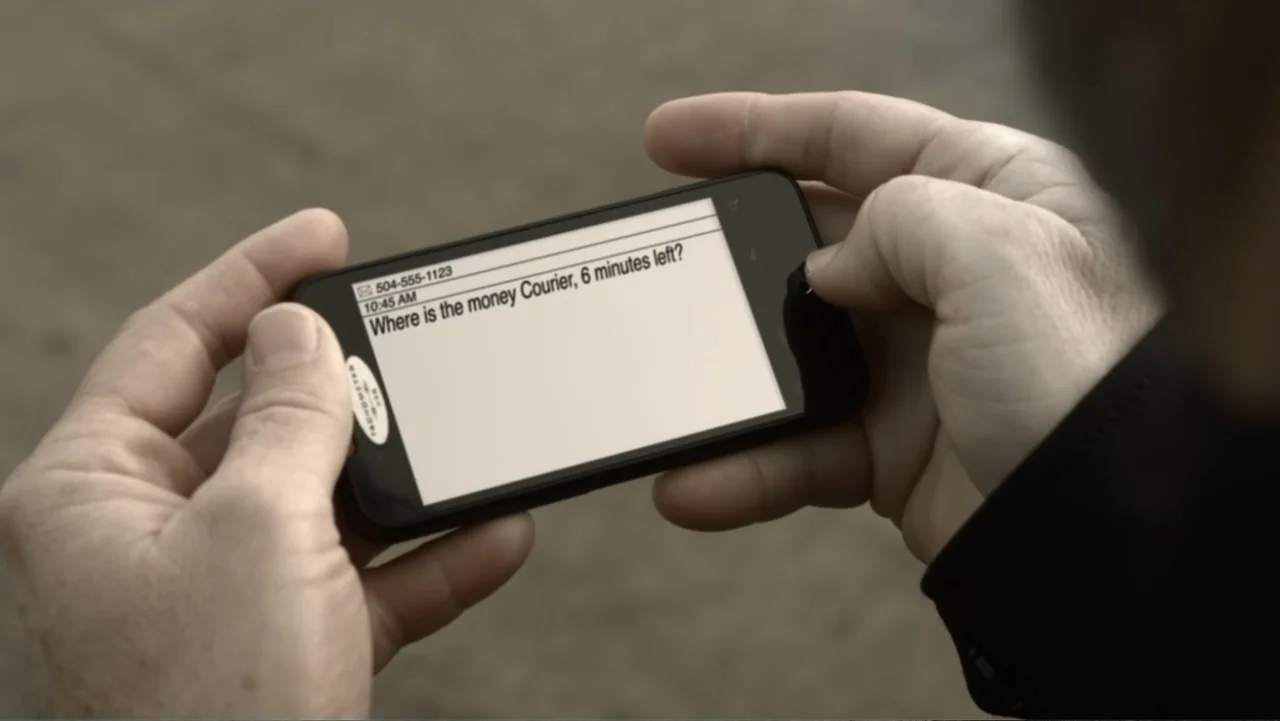 The Messenger (2015) movie still: closeup — A pair of human hands holds a smartphone displaying a text message outdoors. The screen shows a message…