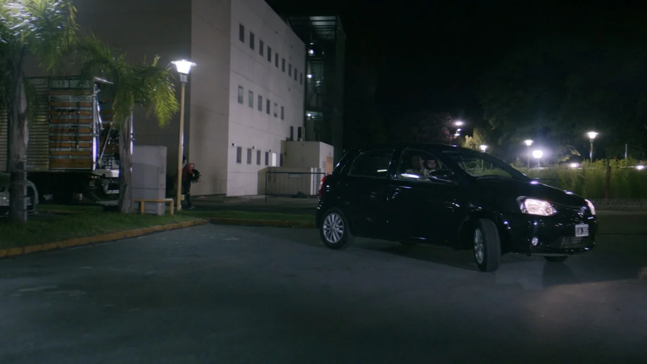 That's Not Cheating (2016) movie still: wide — A nighttime scene in a paved parking lot outside a multi-story office building. Two people sit inside…