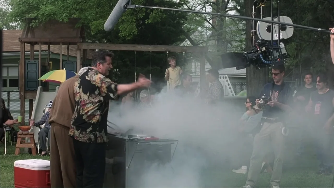 Analyze That (2002) movie still: medium — A backyard setting with a barbecue grill engulfed in thick smoke. A man in a floral shirt gestures towards…