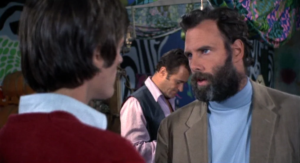 The Trip (1967) movie still: medium — A dimly lit room features patterned fabric wall hangings and blurred furniture pieces. A man in a blue…