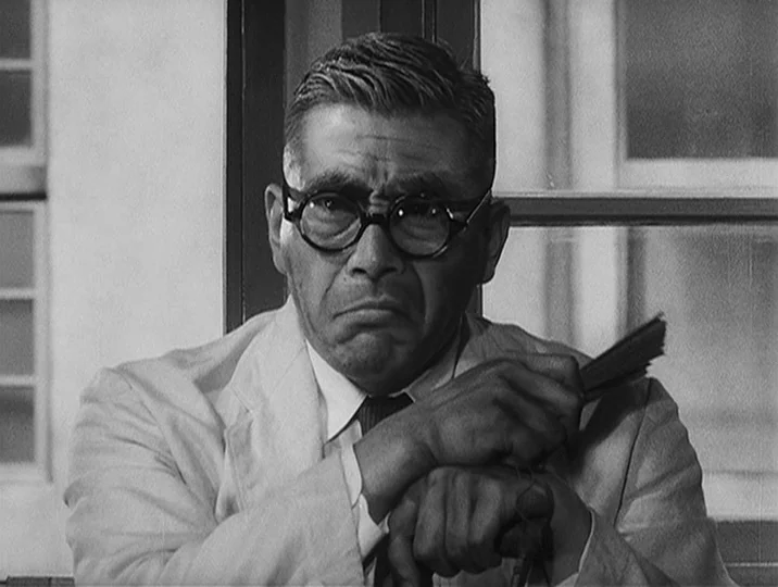 I Live In Fear (1955) movie still: medium — A man in a suit stands indoors with a window visible behind him. The man holds a small folded paper fan…