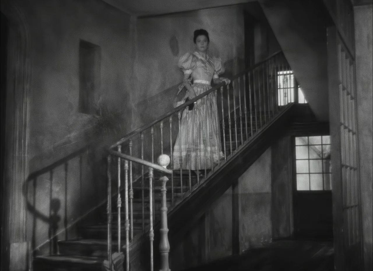 Les Enfants Du Paradis (1945) movie still: fullbody — A woman in a Victorian-style dress stands on a wooden staircase in a dark room. The walls are…