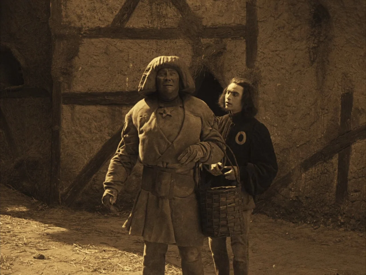 The Golem (1920) movie still: fullbody — A rustic timber-framed building with mud-plastered walls stands behind two people in costume. The…