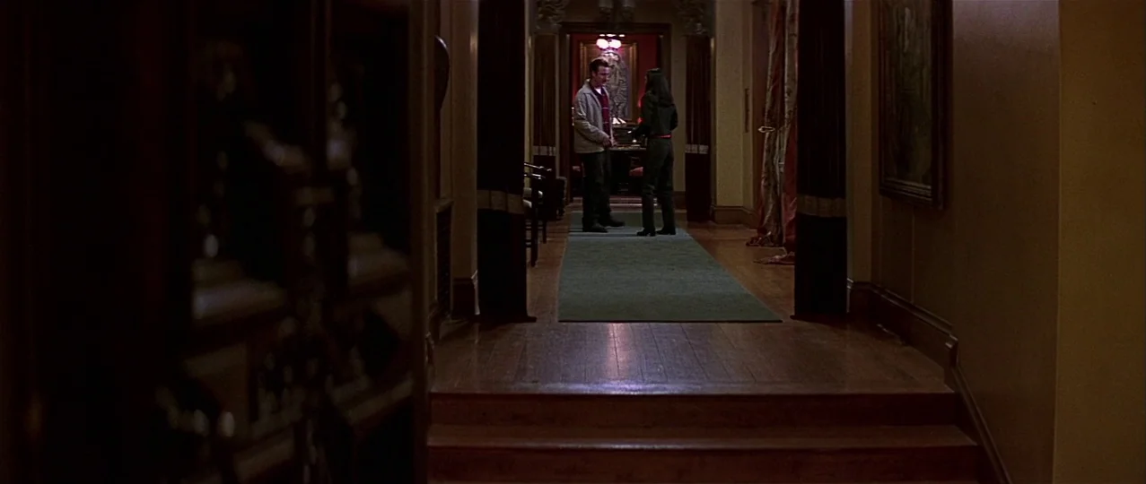 Scream 3 (2000) movie still: wide — A long, shadowed residential hallway features wooden flooring and heavy dark curtains on sides. Two individuals…
