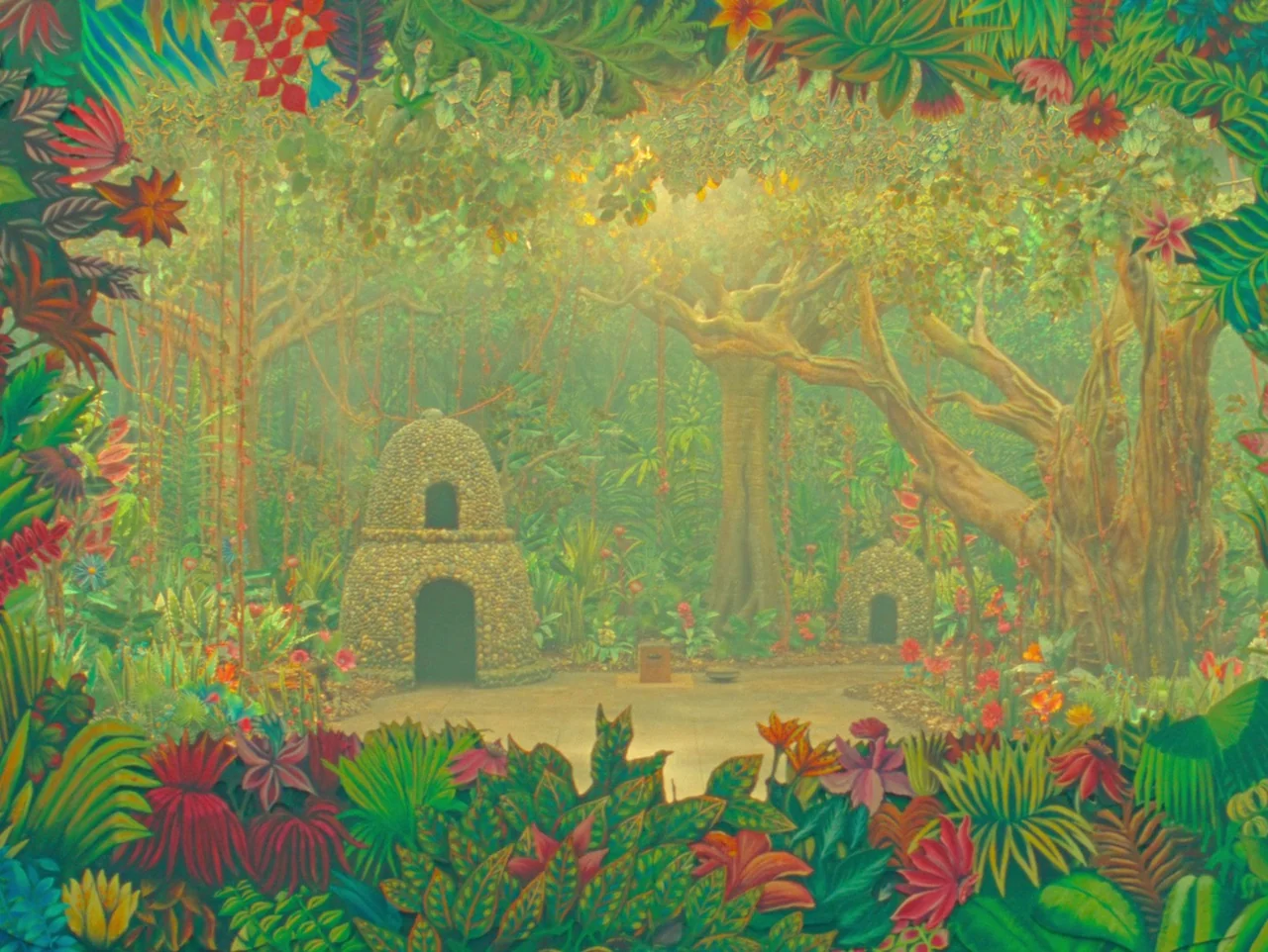 The Wonderful Story of Henry Sugar (2023) movie still: establishing — A dense, vibrant jungle clearing features two small stone-built structures…