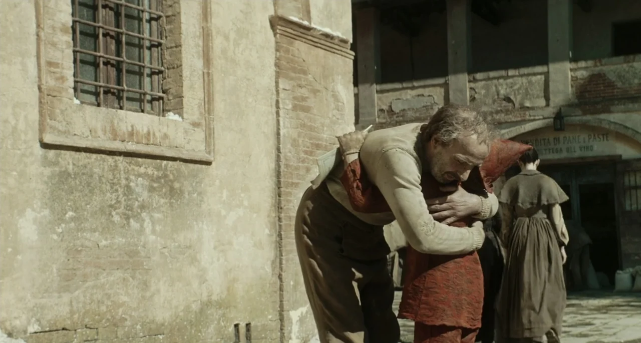 Pinocchio (2022) movie still: medium — A stone building with a barred window sits beside a sunlit courtyard. An older man hugs a person wearing a…