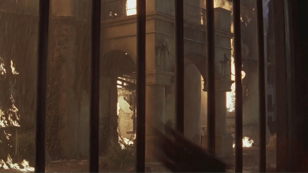The Mask of Zorro (1998) movie still: wide — A stone courtyard with arches is engulfed in bright flames during a heavy rainstorm. Thick black iron…