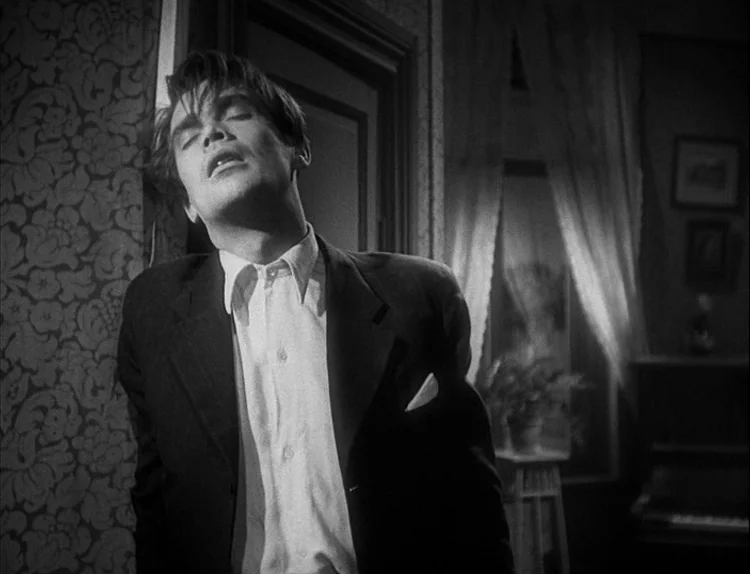 Music in Darkness (1948) movie still: medium — A domestic space with floral patterned wallpaper and a window covered by sheer curtains. A young man…