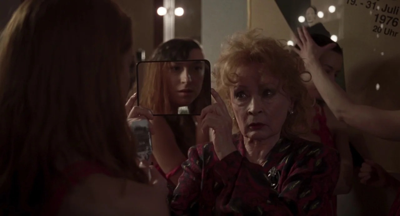 Suspiria (2018) movie still: medium — A cluttered backstage dressing room with illuminated vanity mirrors and vintage event posters. An older woman…
