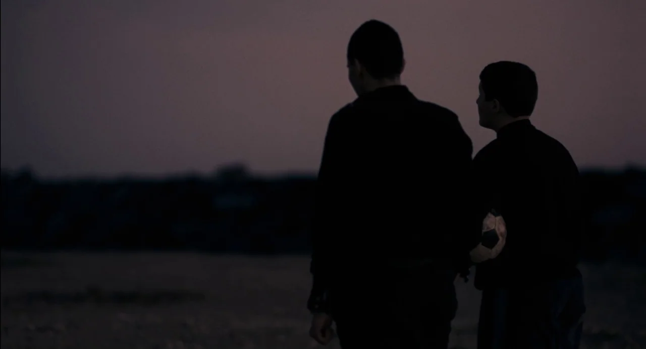 Pasolini (2014) movie still: medium — Two individuals stand in an open outdoor area during the late twilight hours. They watch the horizon while…