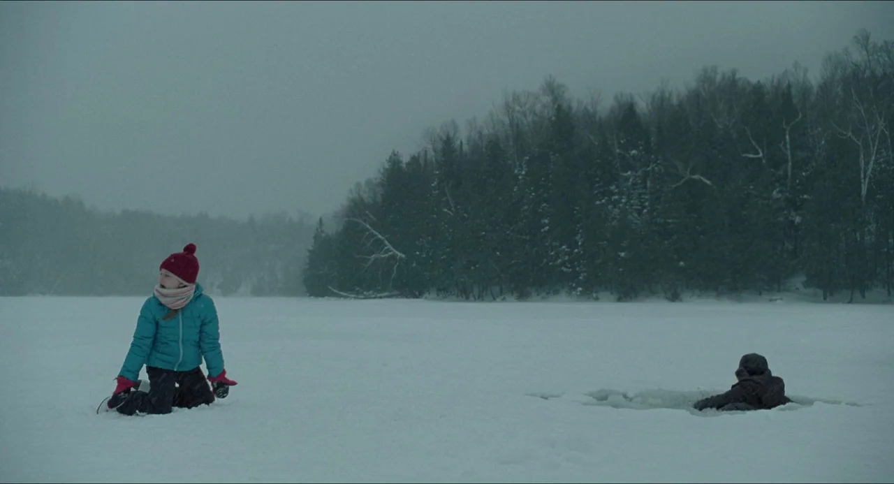 The Lodge (2019) movie still: wide — A vast, white frozen lake surface sits beneath a heavy, overcast grey sky. A girl in a bright blue jacket…