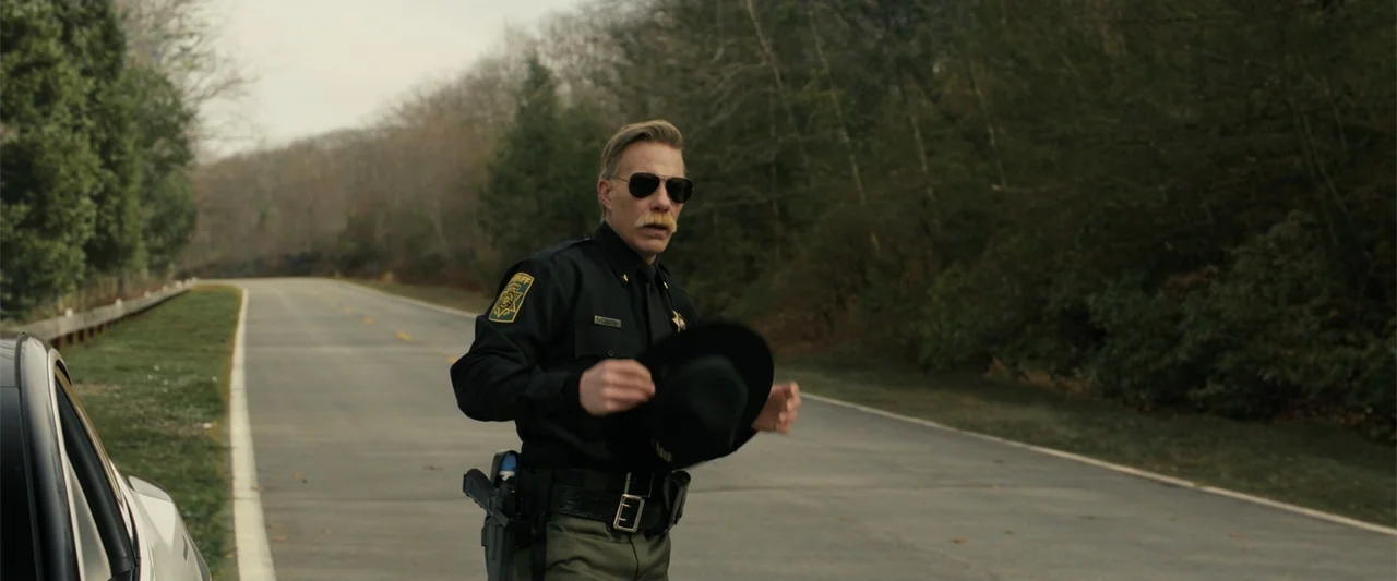 Companion (2025) movie still: medium — A man in a sheriff uniform stands on an empty asphalt road bordered by trees. He holds a black hat in his…