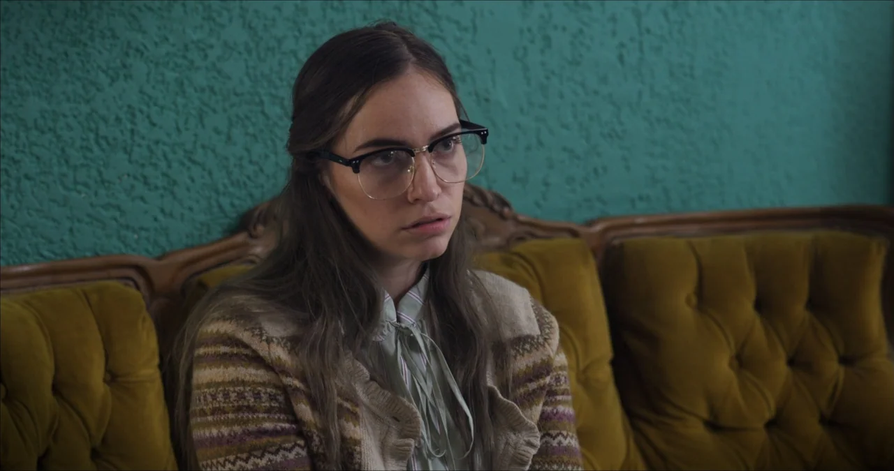 Bendita Suegra (2023) movie still: medium — A 1970s interior with a textured teal wall and a tufted olive-colored sofa. A young woman with glasses…