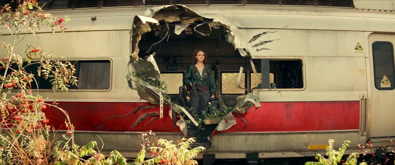 A Quiet Place Part II (2020) movie still: wide — An abandoned passenger train sits outdoors with a large, jagged hole in its side. A young woman in…