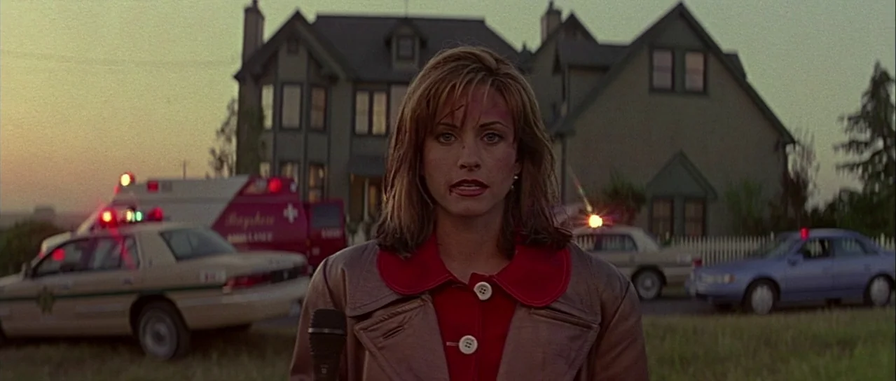 Scream (1996) movie still: medium — A suburban house stands in the background as emergency vehicles are parked on the grass. A female reporter…