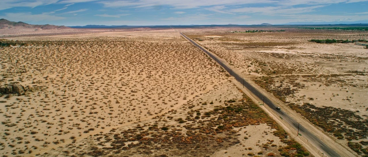 Wish I Was Here (2014) movie still: establishing — A vast, arid desert landscape stretches toward distant mountains under a bright blue sky. A…