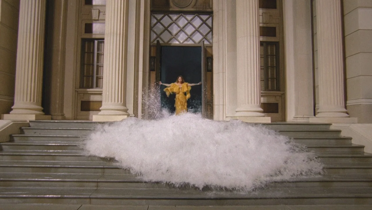 Lemonade (2016) movie still: wide — The front entrance of a classical building with massive columns features an open doorway. A woman in a yellow…
