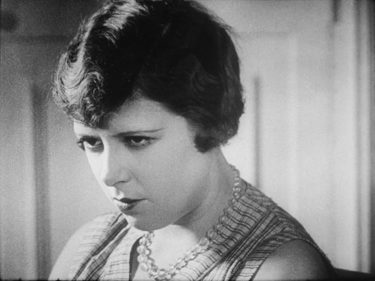 Un Chien Andalou (1929) movie still: closeup — A well-lit indoor space with plain wall panelling as a background. A woman in a patterned dress looks…