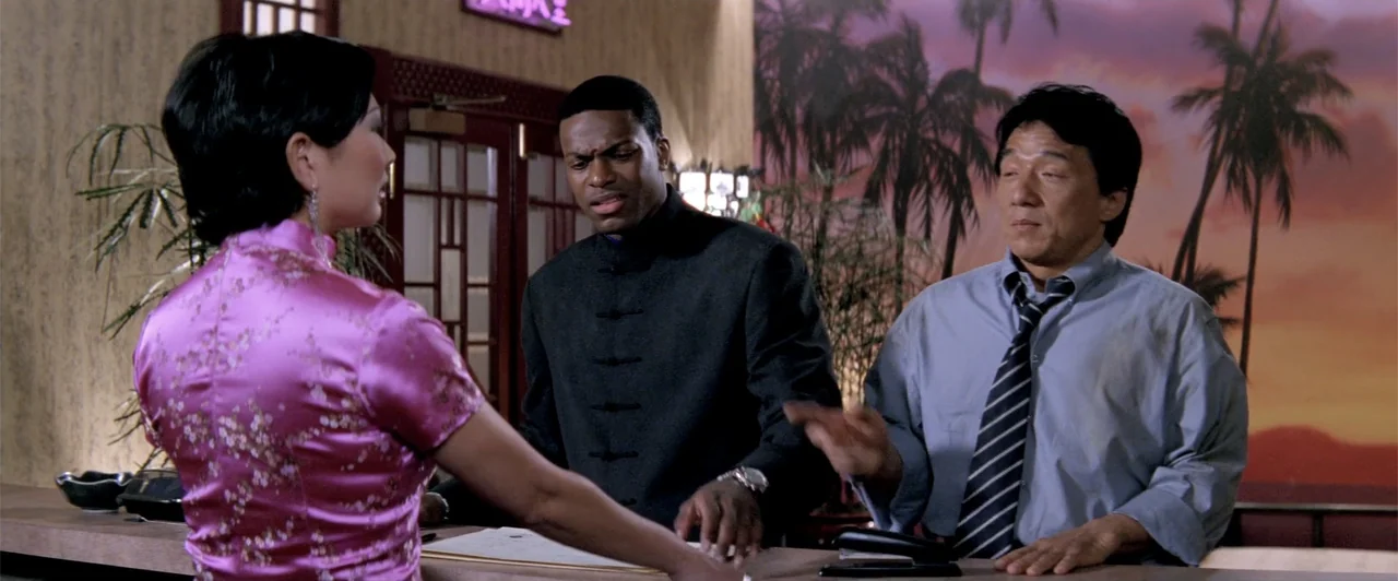 Rush Hour 2 (2001) movie still: medium — A hotel check-in counter features a mural of palm trees at dusk behind guests. A woman in a pink qipao…