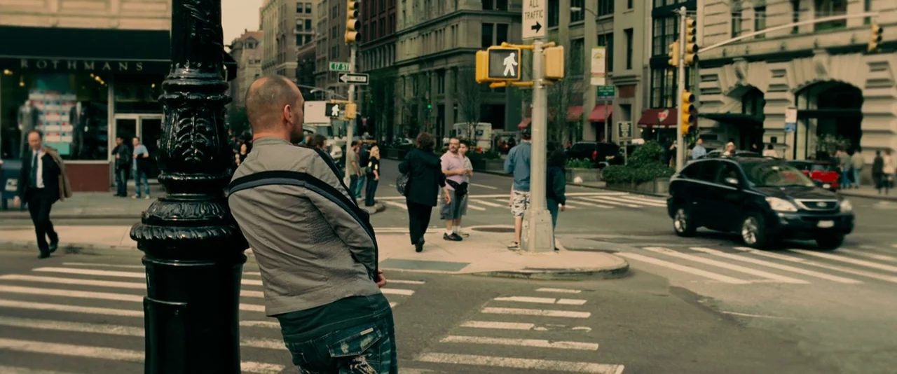 Limitless (2011) movie still: medium — A man leans against a street sign pole at a busy urban crosswalk. Multiple pedestrians walk across the…