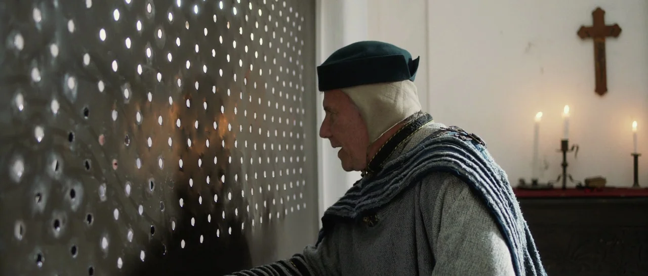 The Little Hours (2017) movie still: medium — A man in medieval attire peers through a perforated metal screen at a partition. A wooden crucifix…