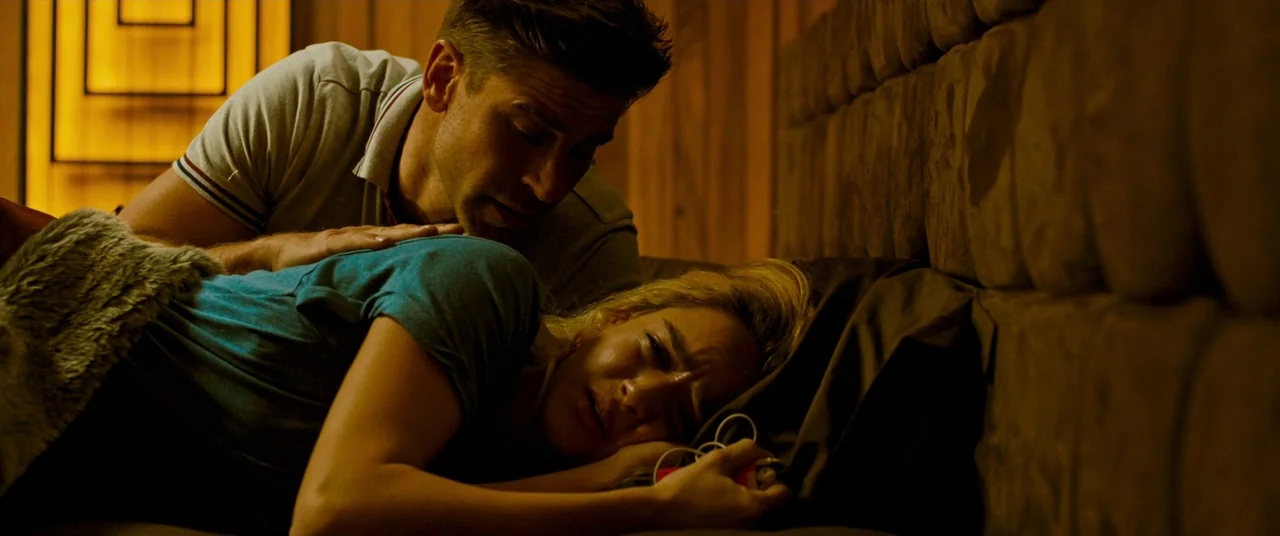 Revenge (2017) movie still: medium — A modern bedroom features wood-paneled walls and a tufted fabric headboard. A man leans over a woman as she…
