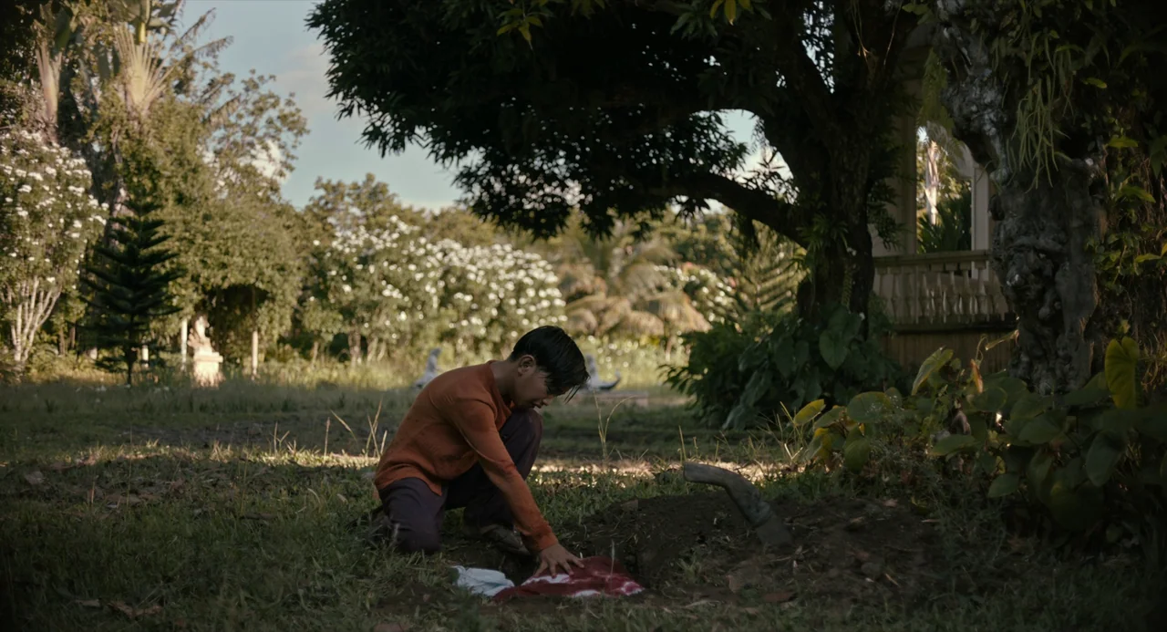 In My Mother’s Skin (2023) movie still: fullbody — An overgrown yard with a large leafy tree and flowering shrubs in the distance. A teenager in an…
