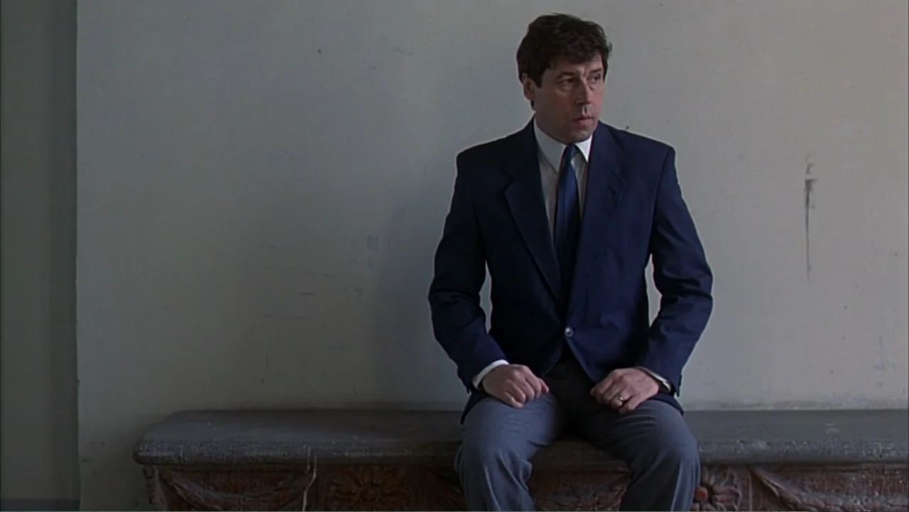 Citizen X (1995) movie still: medium — A man in a business suit sits on a long stone bench against a blank wall. He looks toward the right side of…