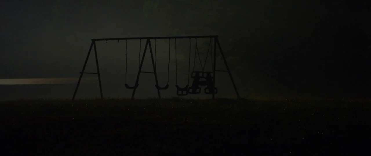 The Strangers: Prey at Night (2018) movie still: wide — A dark, misty field at night contains a metal swing set silhouetted against fog. The swings…