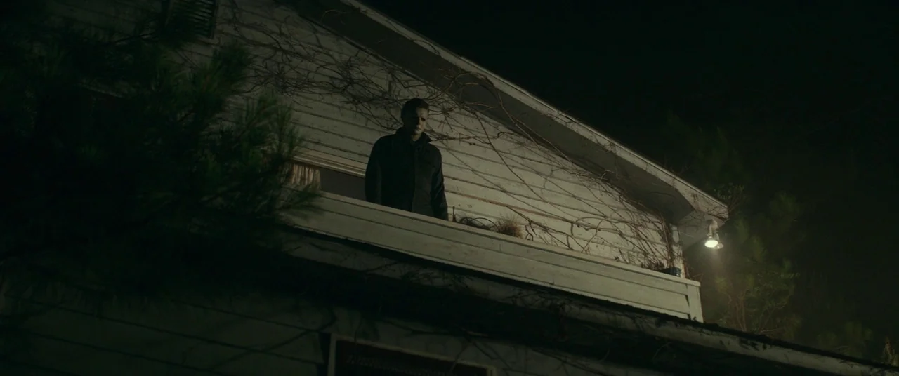 Halloween (2018) (2018) movie still: wide — A white wooden house exterior is covered in dry, dead vines at night. A man in a mask stands on an…